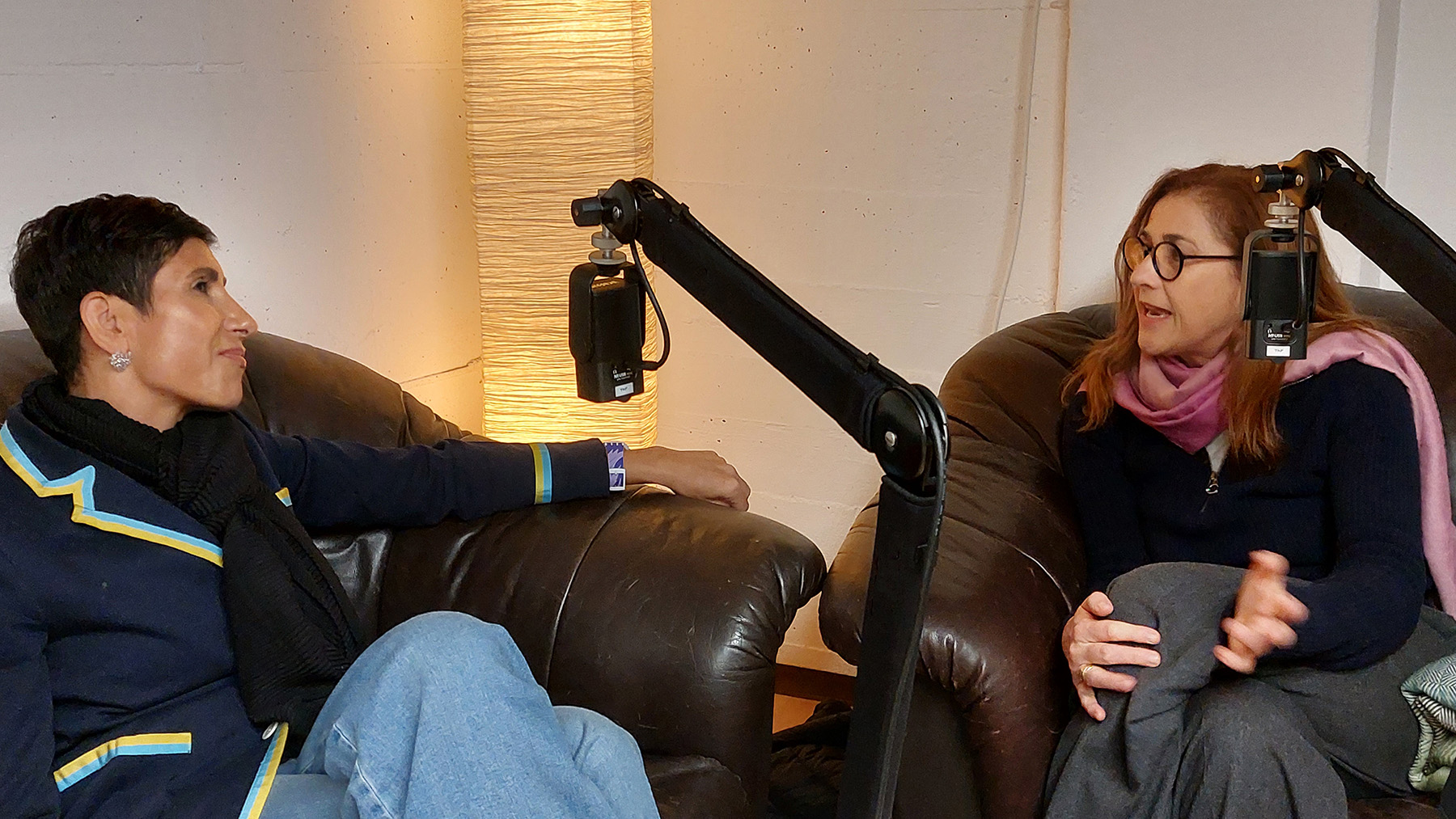 Foto von der Podcast-Aufzeichnung: Zwei Frauen sitzen auf einem Ledersofa und einem Ledersessel, Mikrofone hängen an Mikrofonarmen vor ihnen. Die Frau links gestikuliert mit den Händen und redet, die andere hört ihr ein bisschen skeptisch aber wohlwollend zu