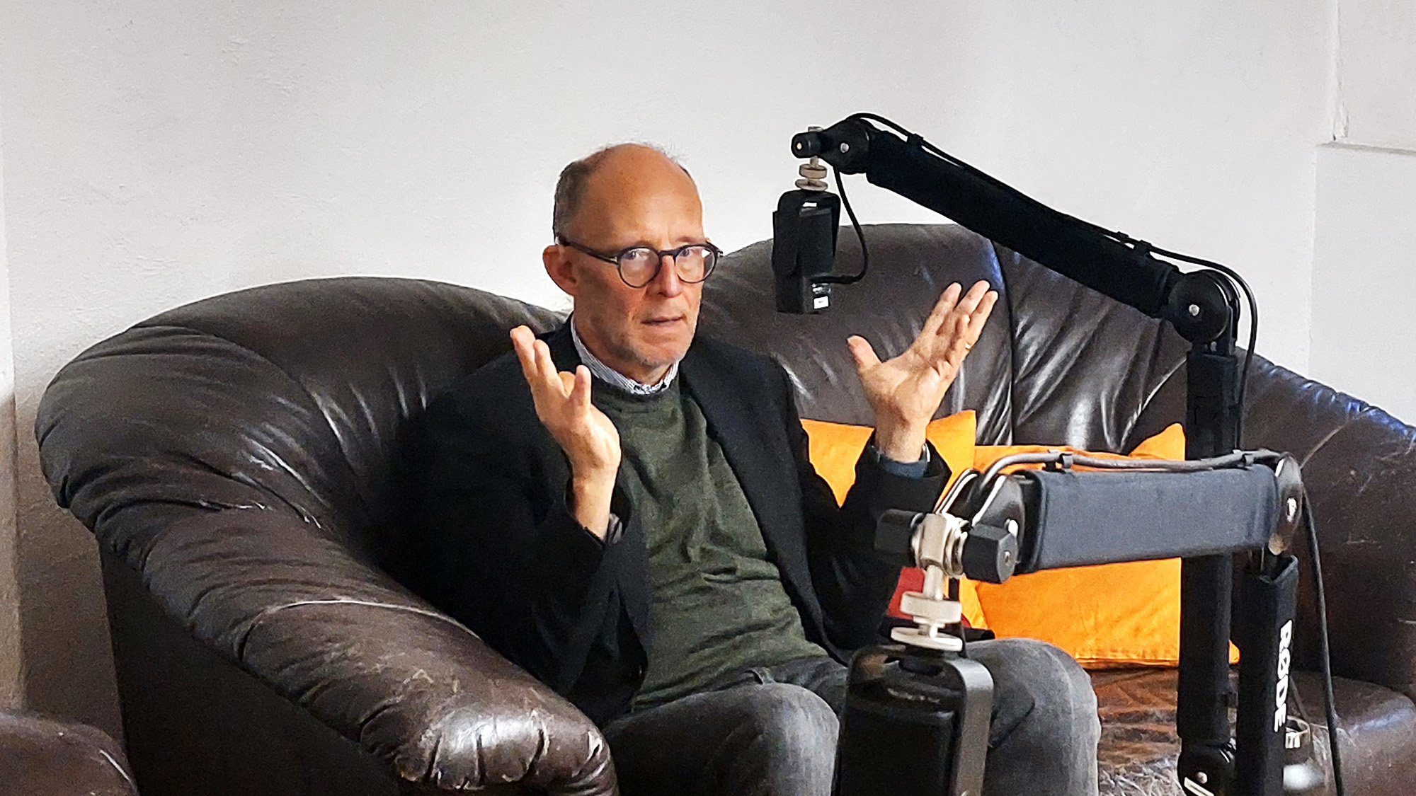 Ralph Kunz sitzt auf einem braunen Sofa (mit orangen Kissen) und gestikuliert mit den Händen, vor ihm – etwa auf Augenhöhe – ein Mikrofon an einem Mikrofonarm. Im Vordergrund sieht man noch einen Teil des Mikrophons von Dorothea Lüddeckens, sie ist aber nicht zu sehen.