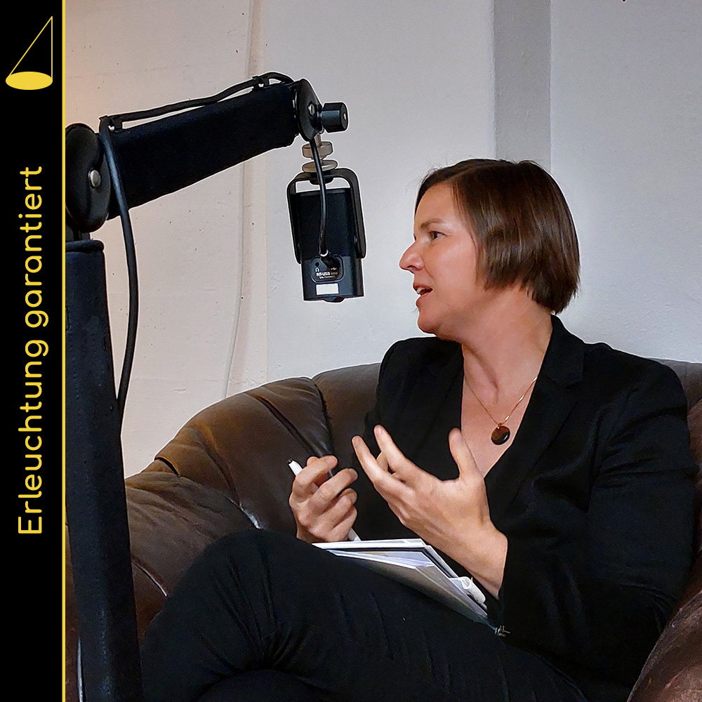 Eine Person mit halblangen Haaren sitzt in einem braunen Ledersessel, blickt und spricht nach links (das Gegenüber ist im Bild nicht zu sehen) und gestikuliert mit den Händen. Vor ihr hängt ein Mikrofon (an einem Mikrofonarm), im Hintergrund eine weisse Wand.