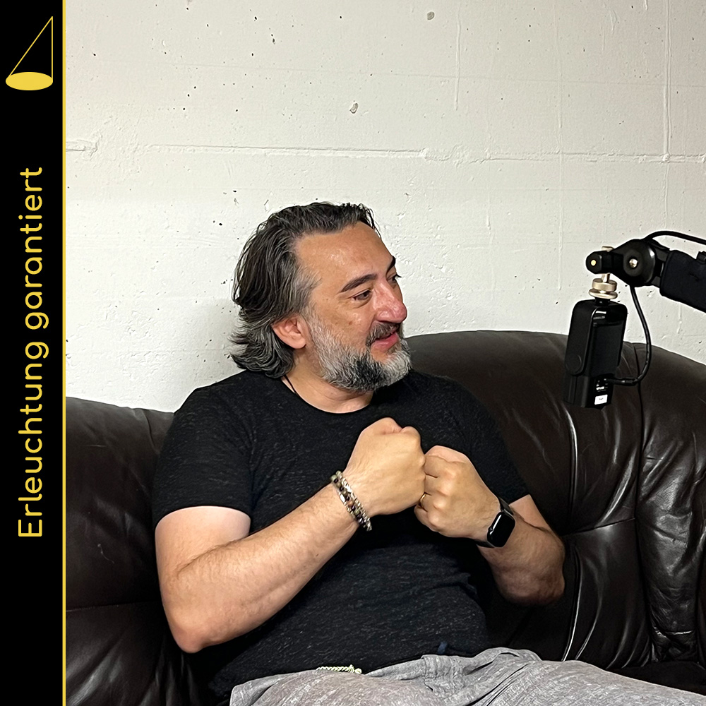Bildbeschreibung: Eine Person mit knapp schulterlangen Haaren und einem Bart (mit weissen und schwarzen Haaren) sitzt auf einem braunen Ledersofa, blickt freundlich nach rechts (das Gegenüber ist im Bild nicht zu sehen) und hält gestisch sein zur Faust geballten Hände zusammen. Vor der Person hängt ein Mikrofon (an einem Mikrofonarm), im Hintergrund eine weisse Wand.