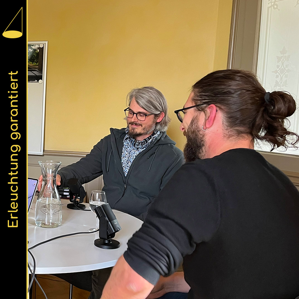 Zwei Personen sitzen nebeneinander an einem runden, weissen Sitzungstisch (man sieht nur die Hälfte, an der sie sitzen), vor ihnen stehen Mikrophone, ein Wasserkrug und Gläser auf dem Tisch. Sie lächeln und blicken in die Mitte des Tischs.