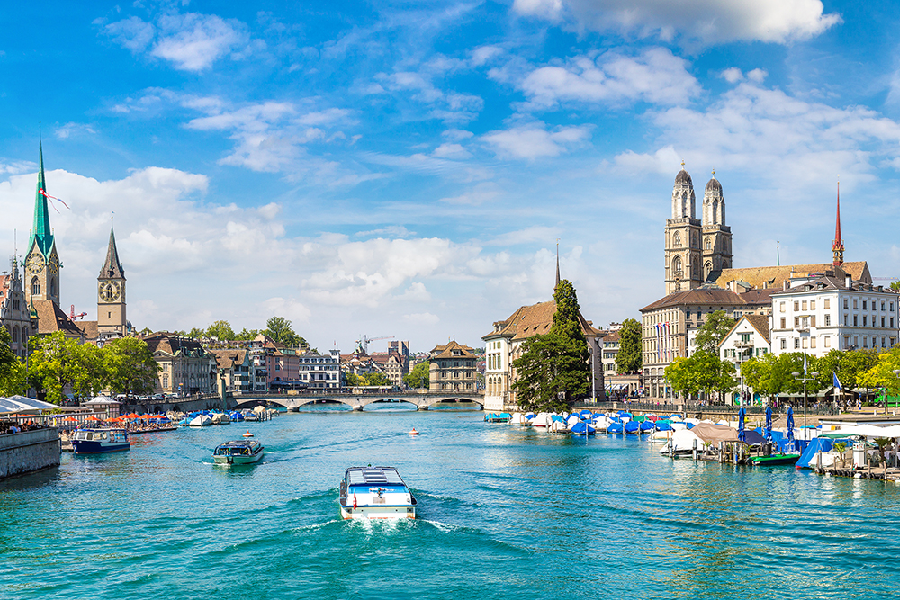 Zürich im Sommer, Blick vom See in Richtung Limmat/Altstadt, im Vordergrund Boote, rechts das Grossmünster, links das Fraumünster; azurblaues Wasser, leuchtend blauer Himmel mit eine paar kleinen Wolken