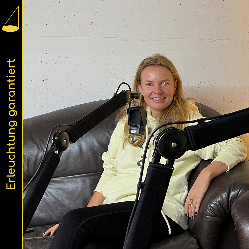 Bildbeschreibung: Eine Person mit langen blonden Haaren und pastellgelbem Pullover sitzt leicht nach vorne gebeugt in einem braunen Ledersofa und blickt lächelnd in die Kamera, vor ihr hängt ein Mikrofon an einem Mikrofonarm. Ein weiterer Mikrofonarm ragt im Vordergrund durchs Bild, Person und Mikrofon sind aber nicht zu sehen.