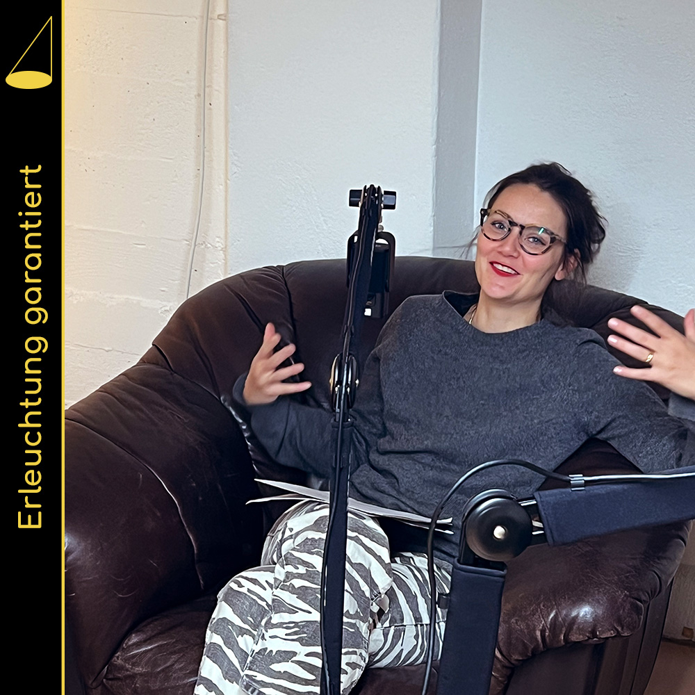 Bildbeschreibung: Eine Person sitzt zurückgelehnt in einem Ledersessel, blickt in die Kamera, lächelt und gestikuliert mit den Armen. Vor ihr ein Mikrofon.