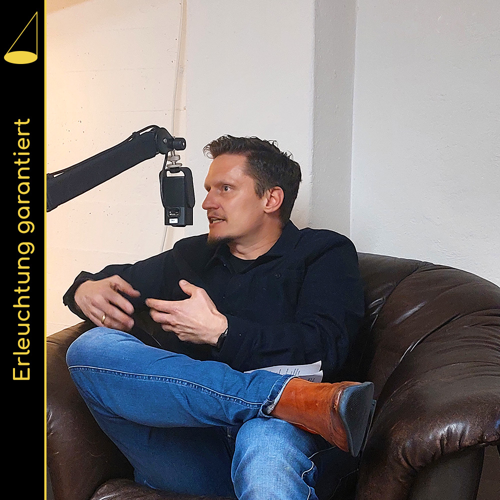 Eine Mann in Jeans sitzt mit verschränkten Beinen in einem braunen Ledersessel und gestikuliet mit beiden Händen. Vor seinem Mund hängt ein Mikrofon an einem Mikrofonarm.