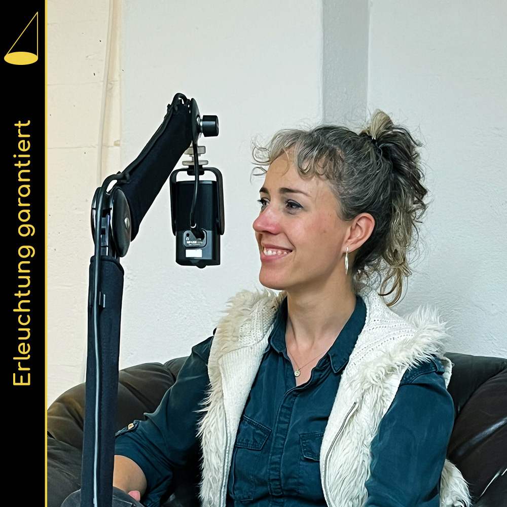 Eine Person mit zusammengebundenen Haaren sitzt in einem braunen Ledersessel, blickt nach links (das Gegenüber ist im Bild nicht zu sehen) und lächelt. Vor ihr hängt ein Mikrofon (an einem Mikrofonarm), im Hintergrund eine weisse Wand.