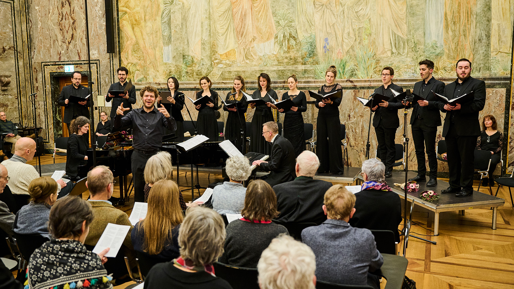 Foto von der Uraufführung der Markuspassion in der Aula der UZH, Blick von hinten auf die Bühne, im Zentrum der zum Publikum gewandte Dirigent, dahinter auf beiden Seiten Flügel und Klavier, noch weiter hinten der Chor. Im Vordergrund sieht man die ersten drei Reihen des Publikums von hinten.