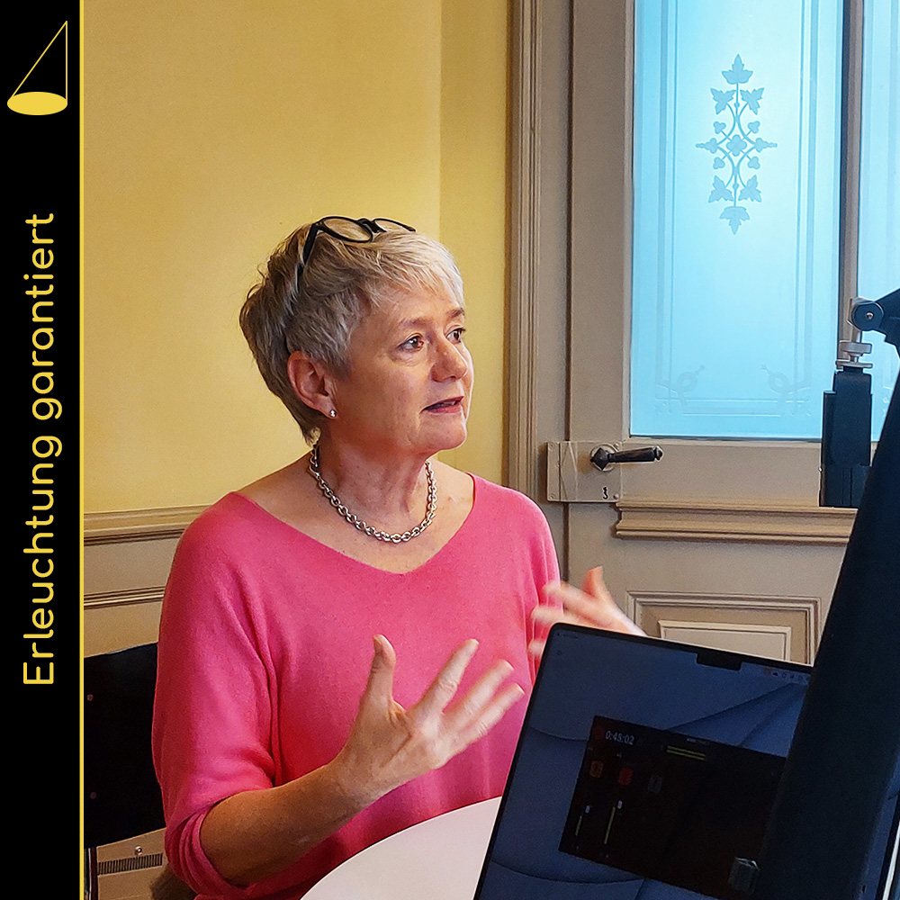 Eine kurzhaarige Frau mit pinkem Oberteil sitzt aufrecht auf einem Stuhl und gestikuliet mit beiden Händen. Im Vordergrund sieht man halbdunkel einen Bildschirm mit eingebledeter Tonaufnahme-Software, im Hintergrund eine gelbe Wand und eine leichte Holztür mit Glas.