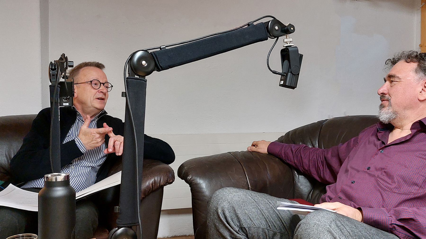 Foto von der Podcastaufnahme mit Rudolf Lutz und Jörg Frey, beide sitzen auf braunen Ledersesseln, im Vordergrund die Mikrofone an Mikrofonarmen. Lutz, links im Bild gestikuliert und blickt zu Frey, rechts im Bild, leicht zurückgelehnt.