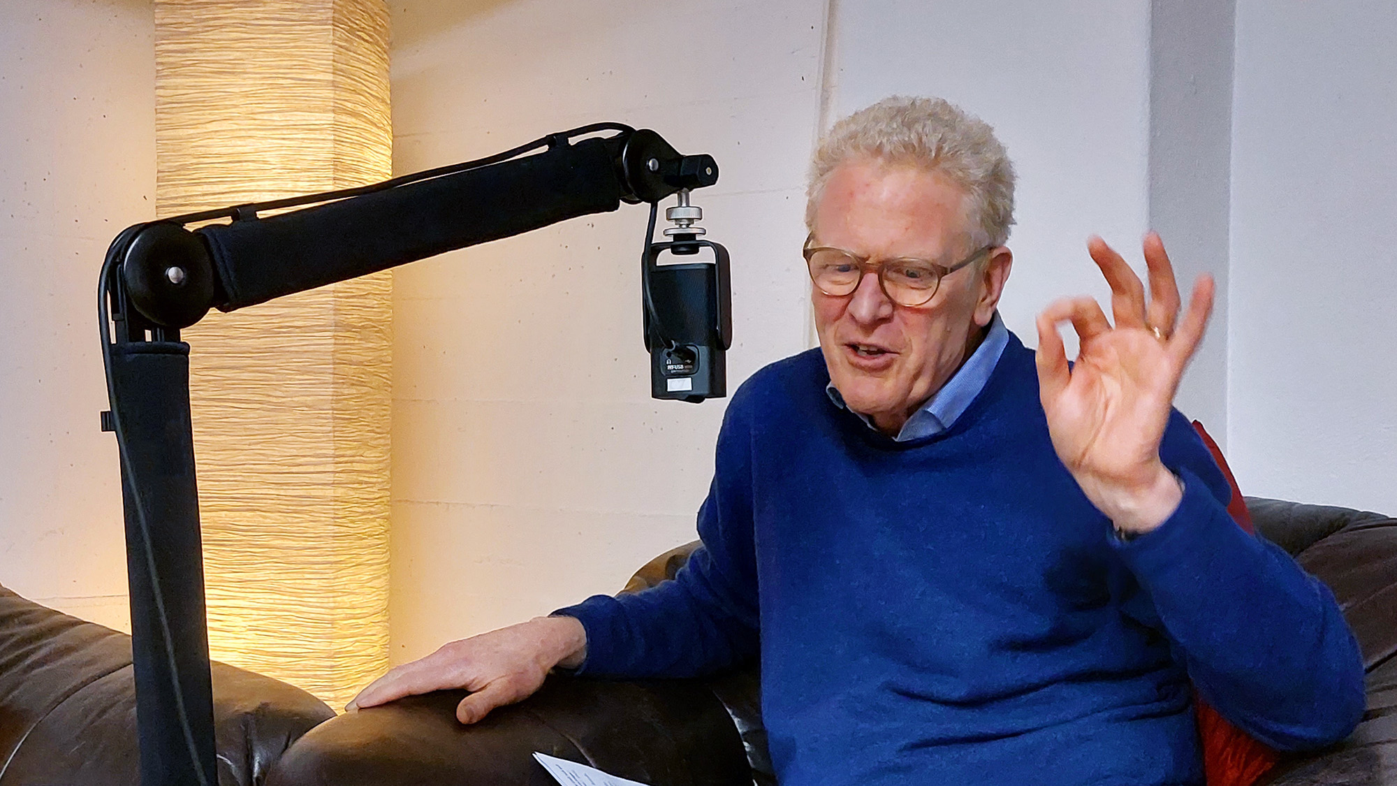 Niklaus Peter sitzt auf einem braunen Sessel und gestikuliert mit der linken Hand (die Spitzen von Zeigefinger und Daumen berühren sich), vor ihm – etwa auf Augenhöhe – ein Mikrofon an einem Mikrofonarm.