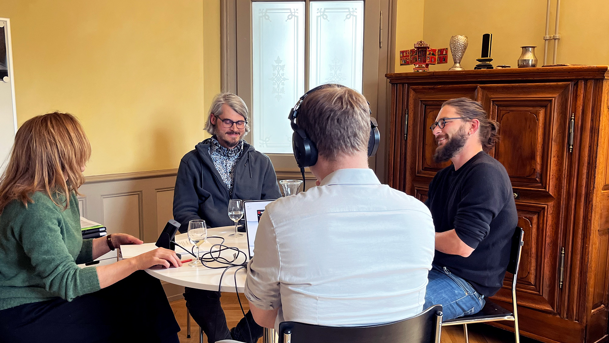 Foto von der Podcast-Aufzeichnung: Vier Personen sitzen um einen kleinen, runden Sitzungstisch, drei Personen haben ein Mikrofon vor sich, von der vierten Person sieht man nur den Rücken (sie hat Kopfhörer auf und einen Computer vor sich). Im Hintergrund eine Türe mit Glaseinsatz und ein antiker Holzschrank.