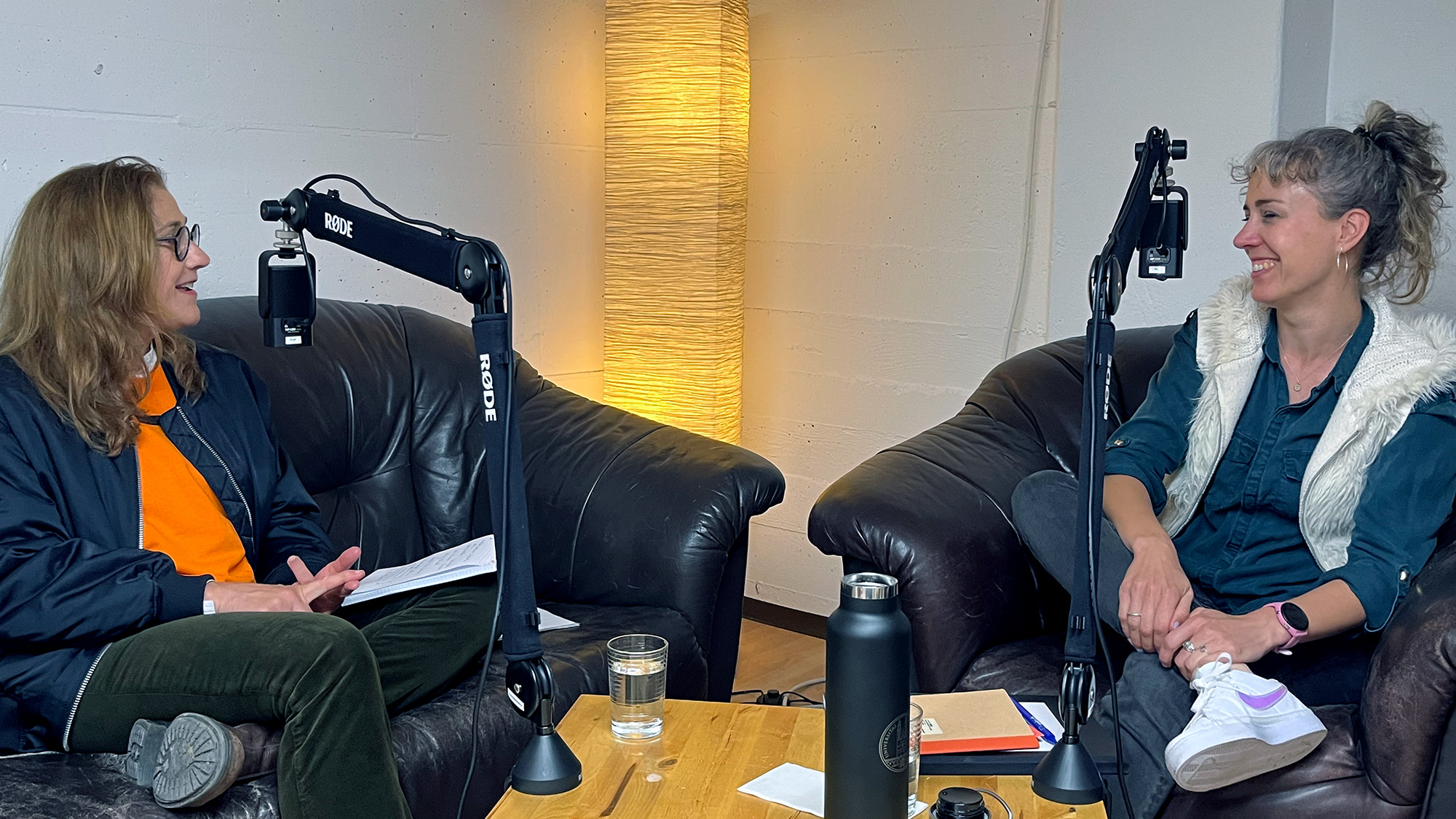Foto von der Podcast-Aufzeichnung: Eine Person sitzt links auf einem dunklen Ledersofa und spricht lächelnd zu ihrem Gegenüber (rechts), dieses sitzt auf einem Sessel derselben Garnitur und lächelt mit zugekniffenen Augen. Im Hintergrund eine weisse Wand mit einer Stehlampe, die das Bild in ein warmes Licht taucht. Vor beiden Personen hängt ein Mikrofon an einem Mikrofonarm.