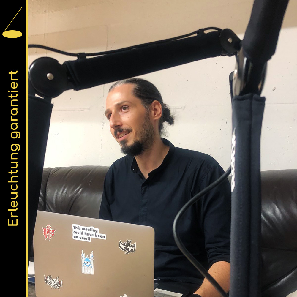 Bildbeschreibung: Eine Person mit mit Bart und zusammengebundenen Haaren sitzt zwischen Mikrofonarmen auf einem Ledersofa, auf den Knien hat sie einen Laptop mit Stickern auf der Rückseite (man sieht nur die Rückseite).