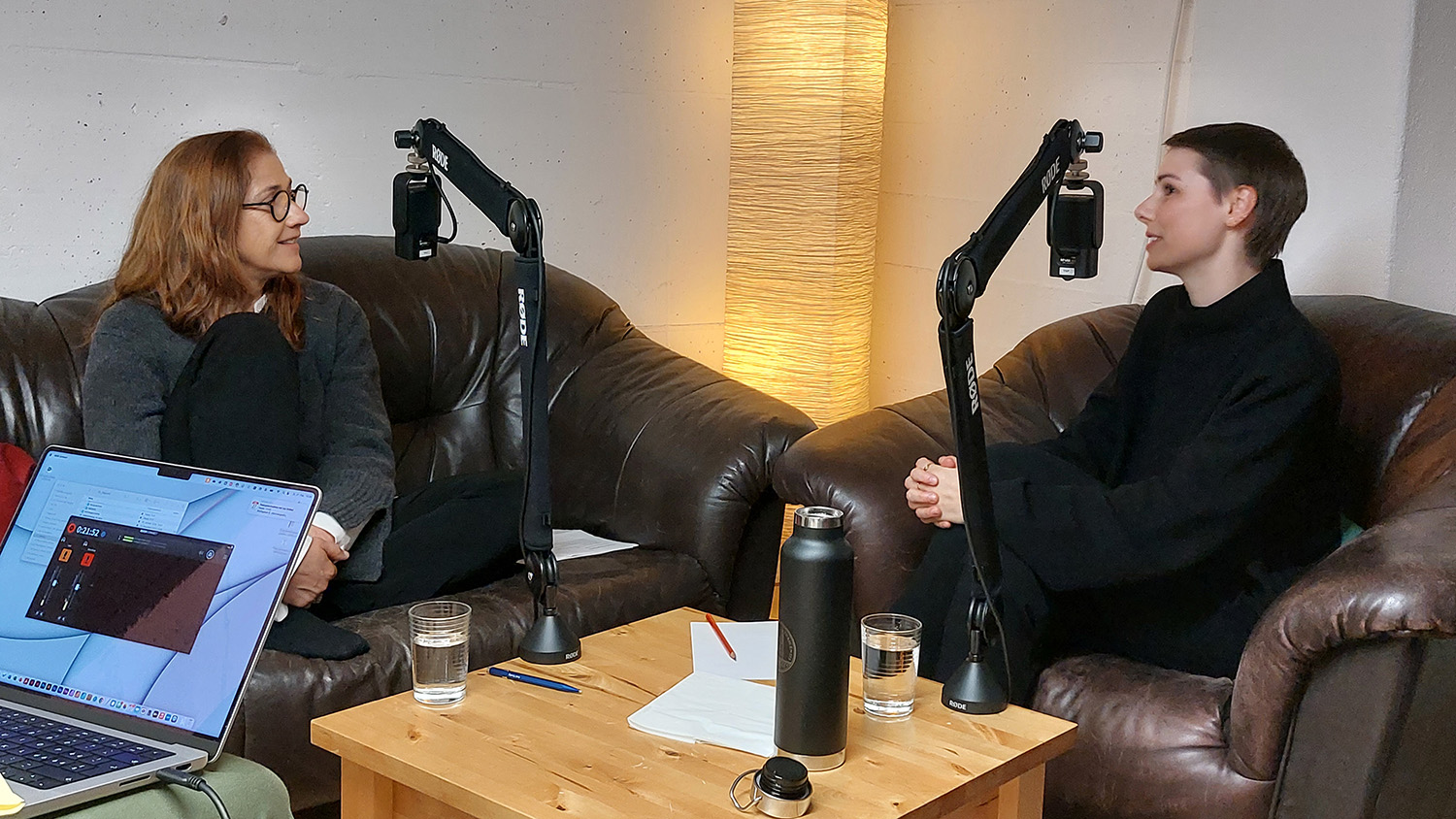 Foto von der Podcast-Aufzeichnung: Ganz links sieht man den Bildschirm eines Computers mit der Aufzeichnung des Audios, weiter hinten sitzen zwei Frauen sich auf Sofa und Sessel gegenüber, vor ihnen je ein Mikrofon an einem Mikrofonarm